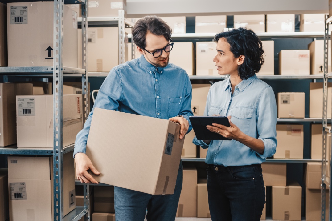 Female Inventory Manager Shows Digital Tablet Information to a Worker Holding Cardboard Box, They Talk and Do Work. In the Background Stock of Parcels with Products Ready for Shipment.